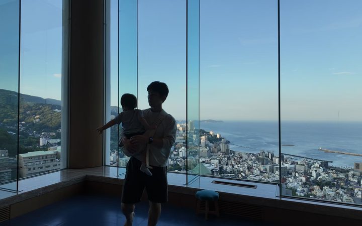 高層階の窓から、父と子で熱海の街と海を眺めるシルエット